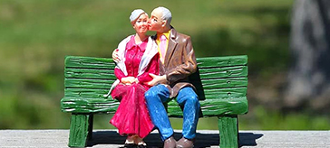 image of a senior couple doll sitting on a green bench in an outdoor setting.