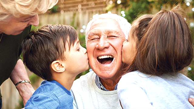 image of a happy male senior laughing while his grandson and grandd-aughter given him a big kiss