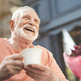 Low angle waist up portrait of delighted mature male sitting at table outside. He is holding cup of hot drink and looking with pleasure and content.