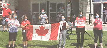 photo of team members from Quinte Gardens Retirement Residence standing at the front entrance and holding up Canada flag wait patiently for the appreciation drive-by on Canada Day.
